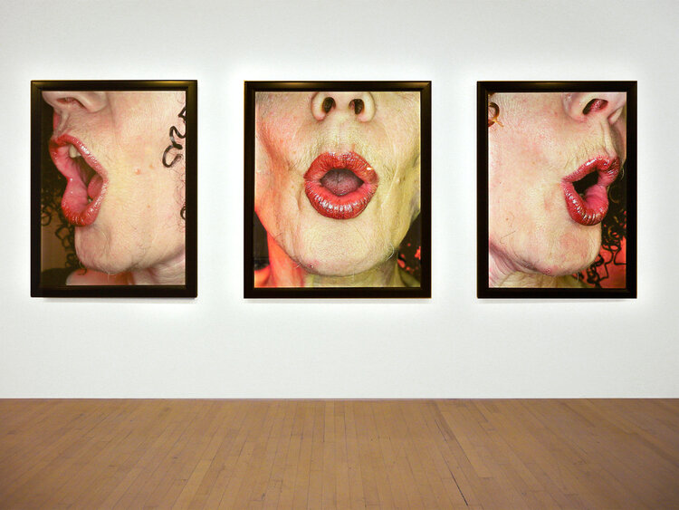 Three close-ups of a woman’s lower face, with lips in an O shape, hung on a gallery wall. She is wearing red lipstick.