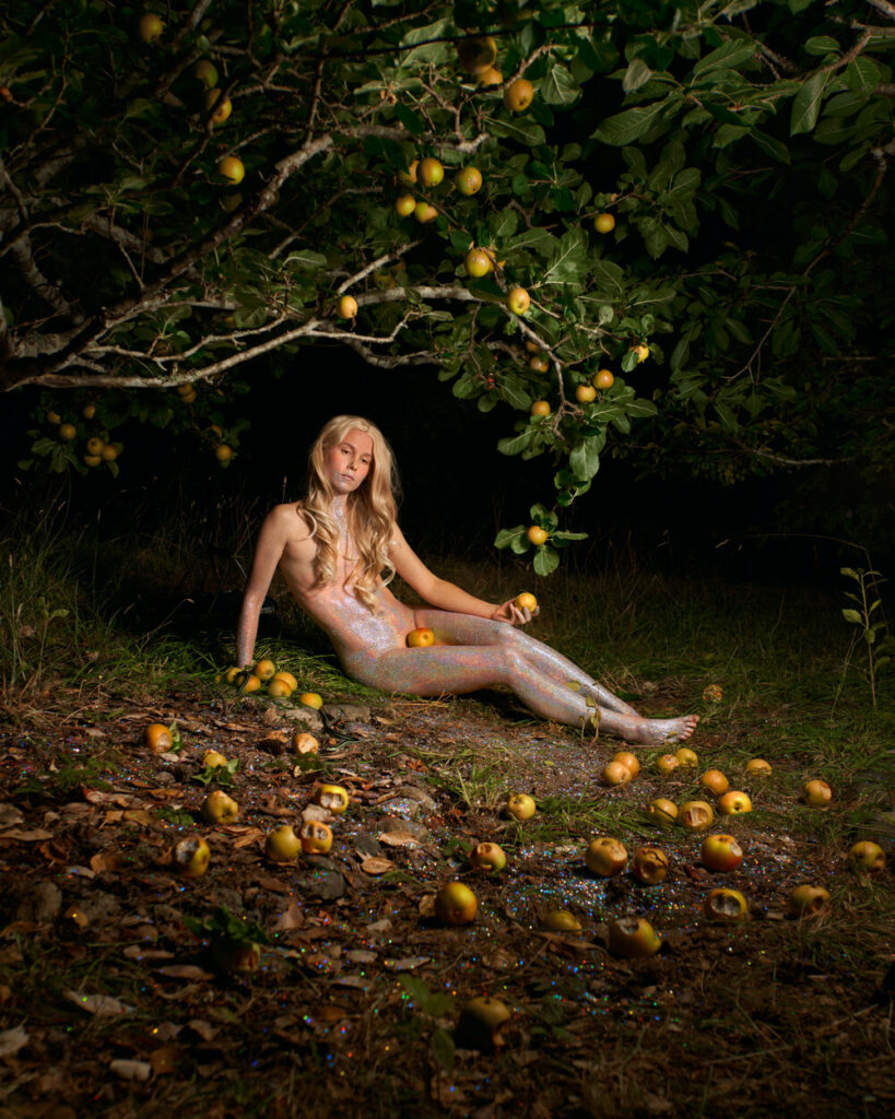 A woman lies under an apple tree at night covered in sparkles. She holds an apple in her hand and one in her lap. Other decaying apples surround her.